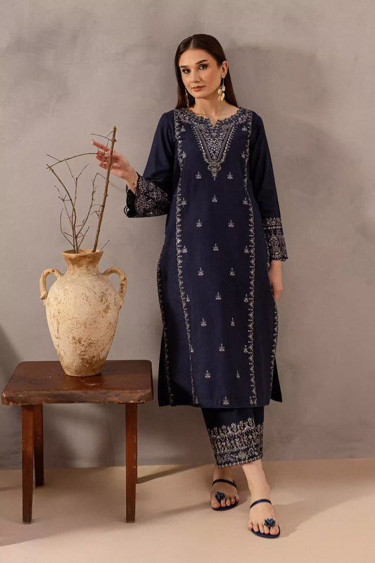 Woman in a navy blue embroidered outfit standing next to a wooden table with a decorative vase.