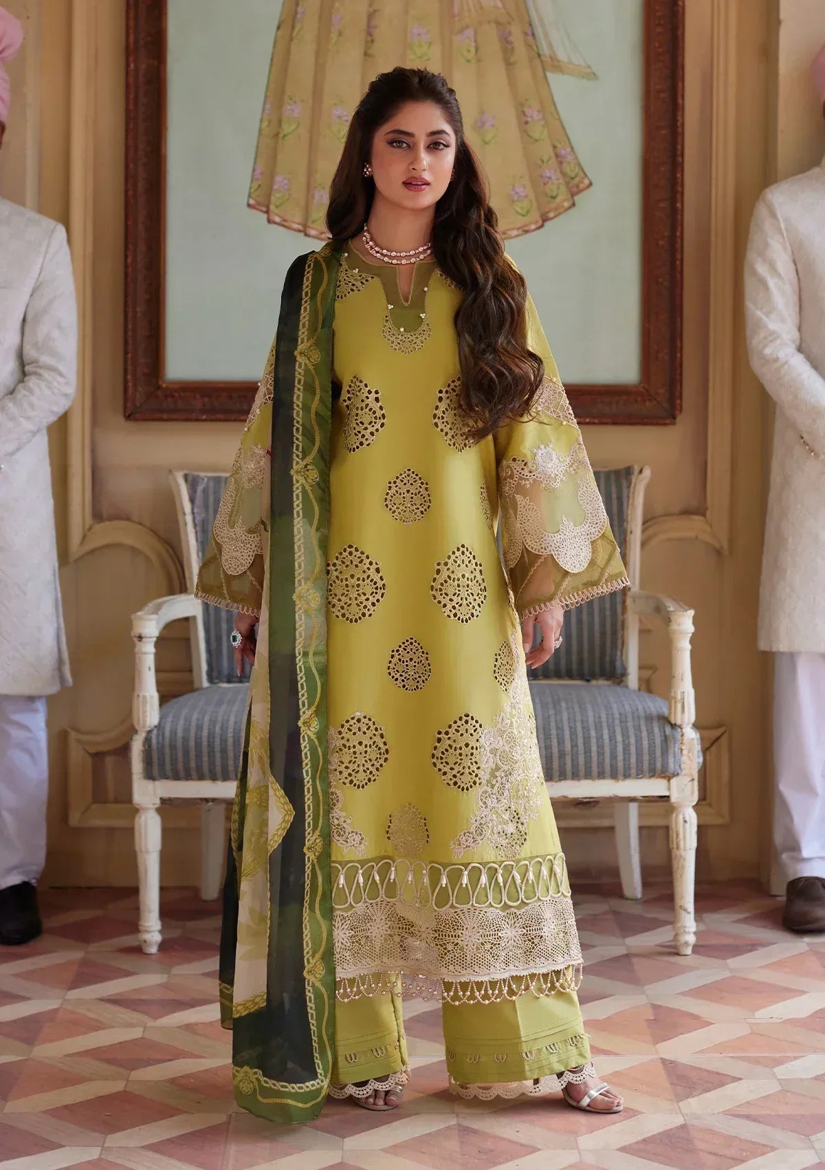 Woman in a yellow and green traditional outfit standing in an elegant room with chairs and a mirror.
