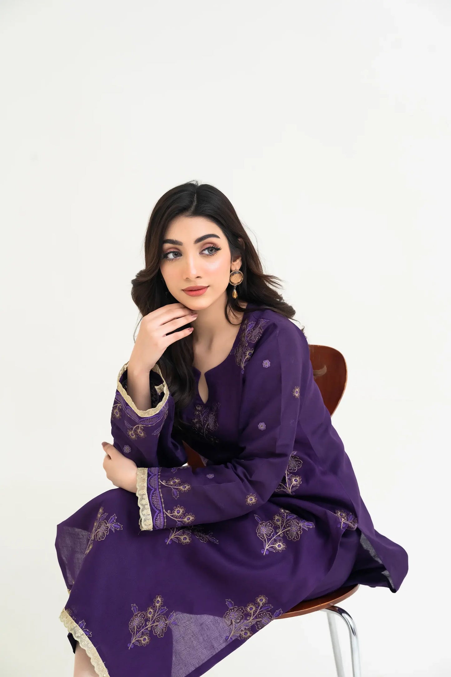 Woman wearing a purple embroidered outfit sitting on a chair against a white background