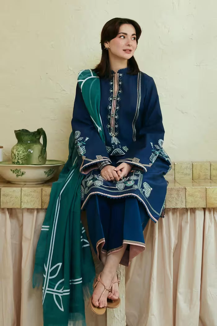 Woman in a blue and green traditional outfit sitting on a ledge with a decorative background.