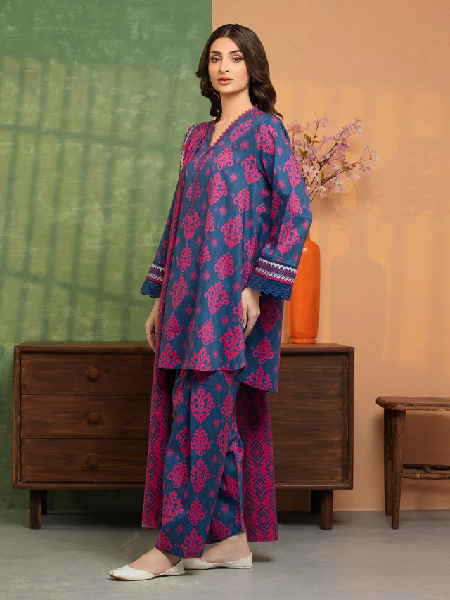 Woman wearing a blue and pink traditional outfit standing in a room with a wooden dresser and vase.