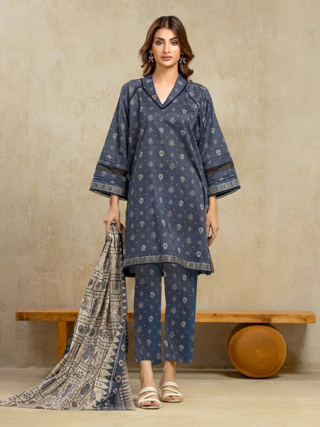 Woman wearing a blue patterned outfit with a scarf, standing against a beige wall.