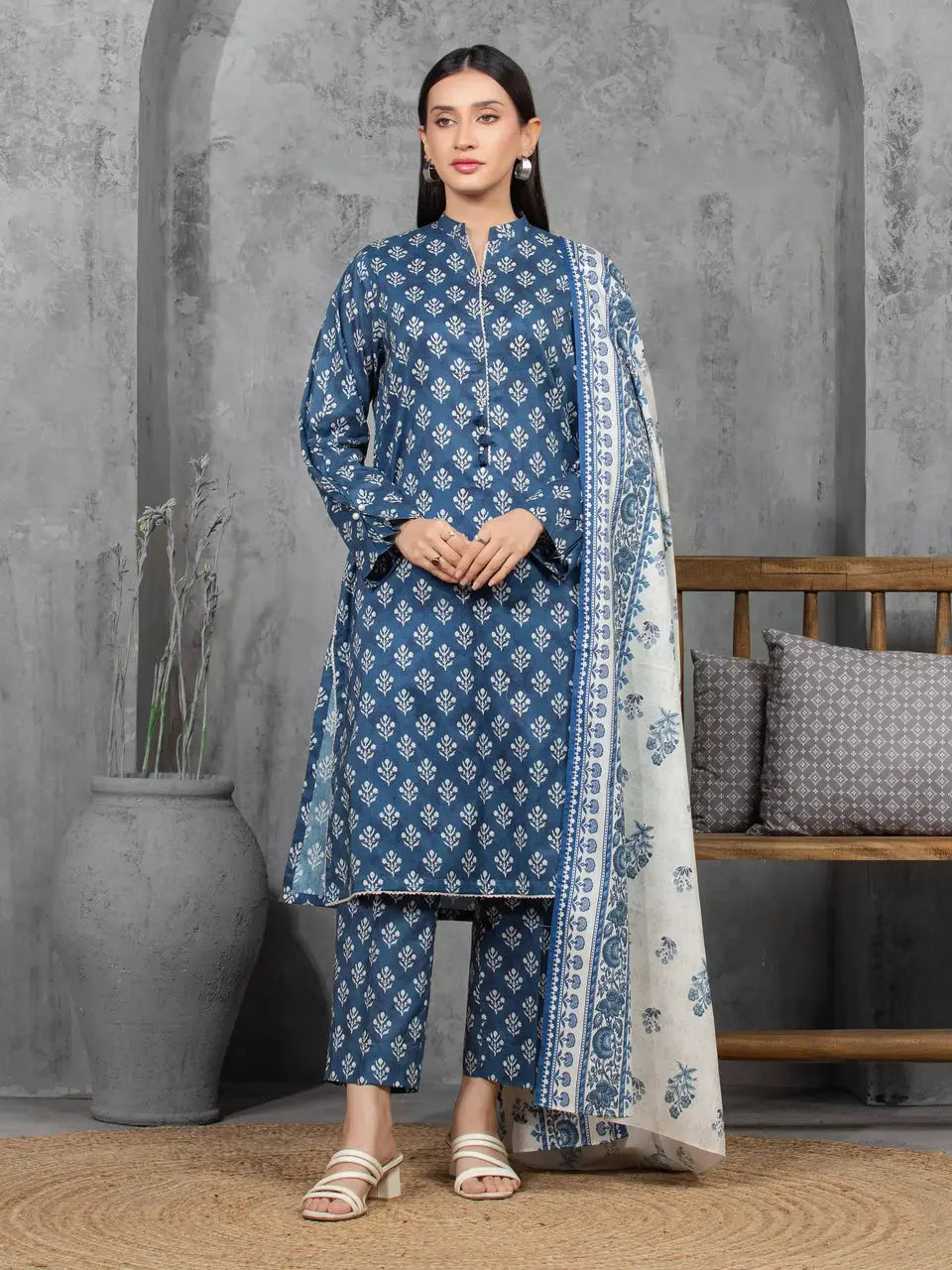 Woman wearing a blue and white patterned traditional outfit with a matching dupatta against a gray wall.