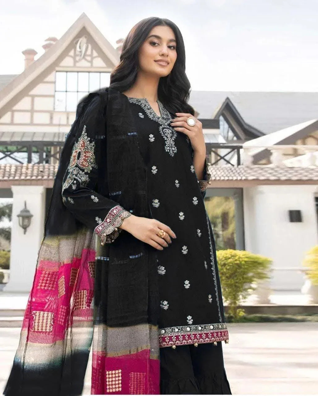 Woman wearing a black embroidered outfit with a colorful dupatta in front of a building.