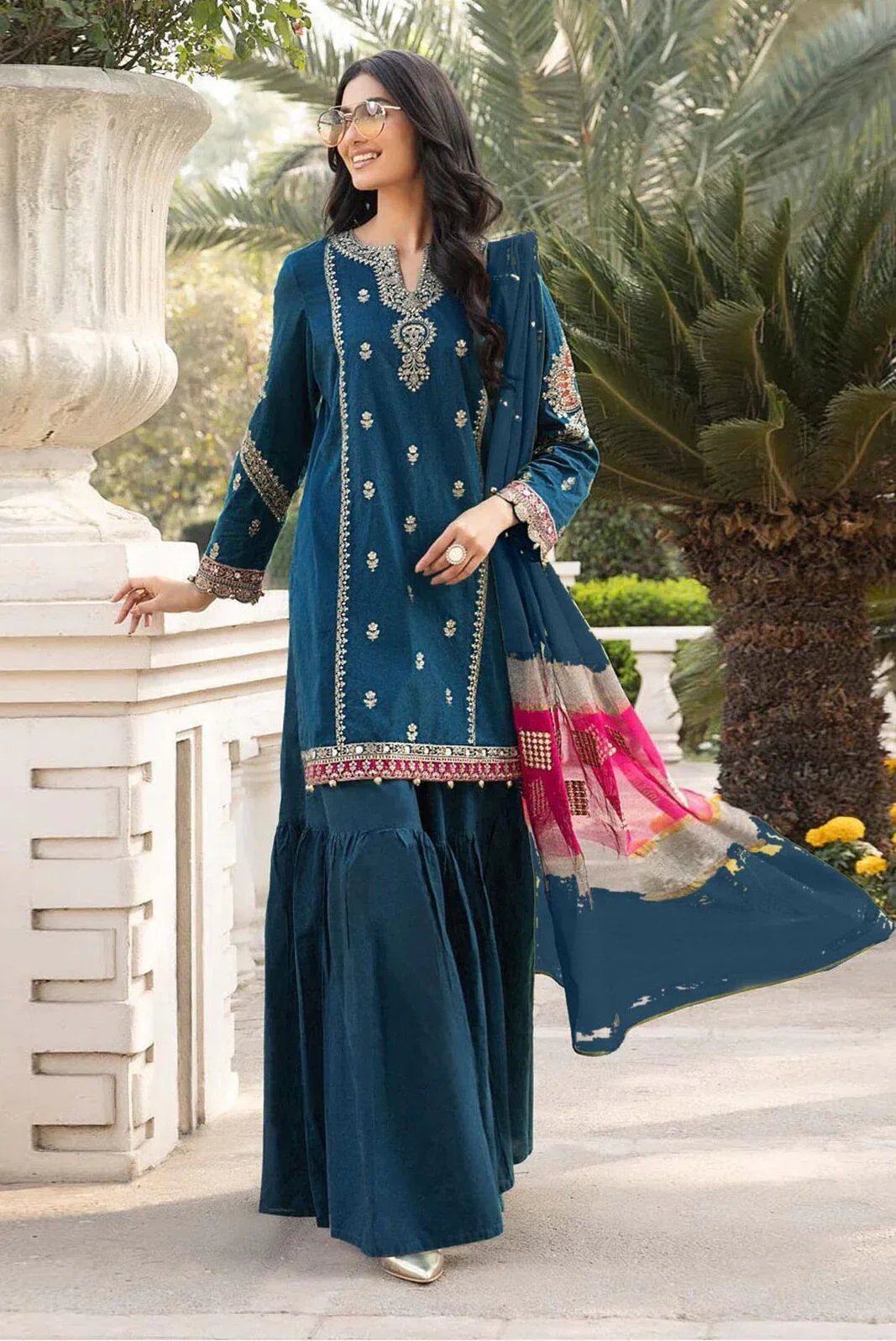 Woman in a teal traditional outfit with pink dupatta standing outdoors.