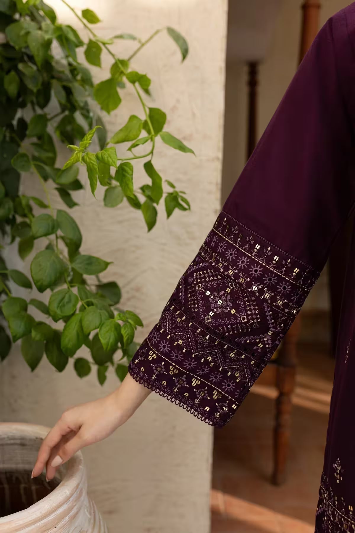 Person wearing a dark purple garment with intricate patterns, standing next to a potted plant.