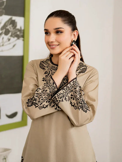 Woman wearing a beige blouse with black embroidery, adjusting an earring indoors.