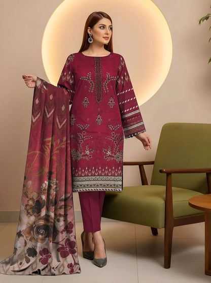 Woman wearing a maroon embroidered outfit with a floral scarf, standing in a room with a green chair and wooden table.
