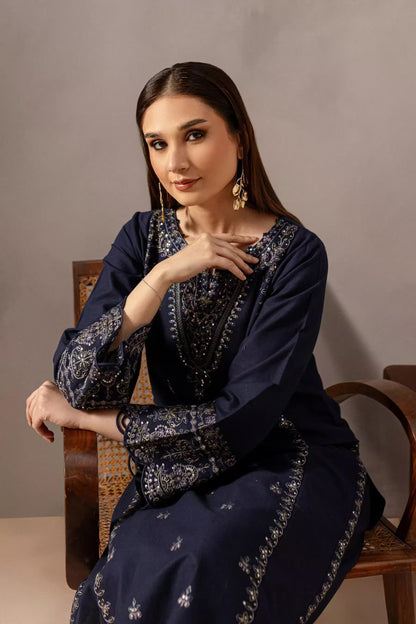 Woman in a dark blue embroidered outfit sitting on a wooden chair against a plain background