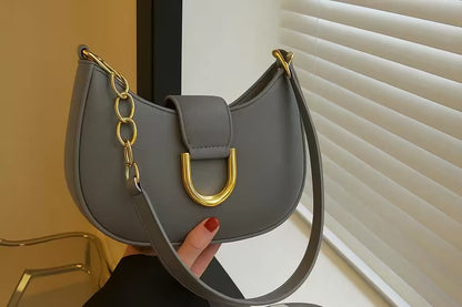 Gray handbag with gold chain and clasp held by a hand against a neutral background