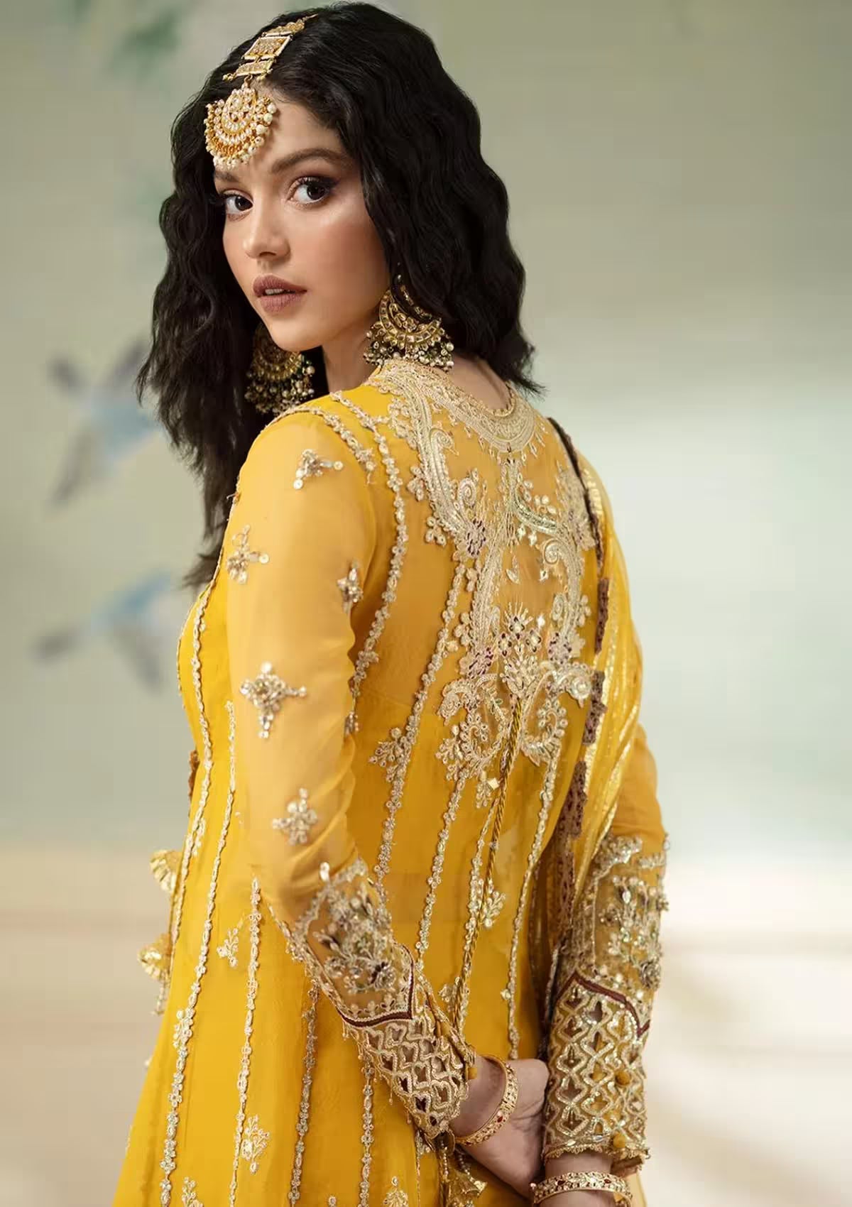 Woman wearing a yellow embroidered traditional outfit with gold jewelry against a neutral background