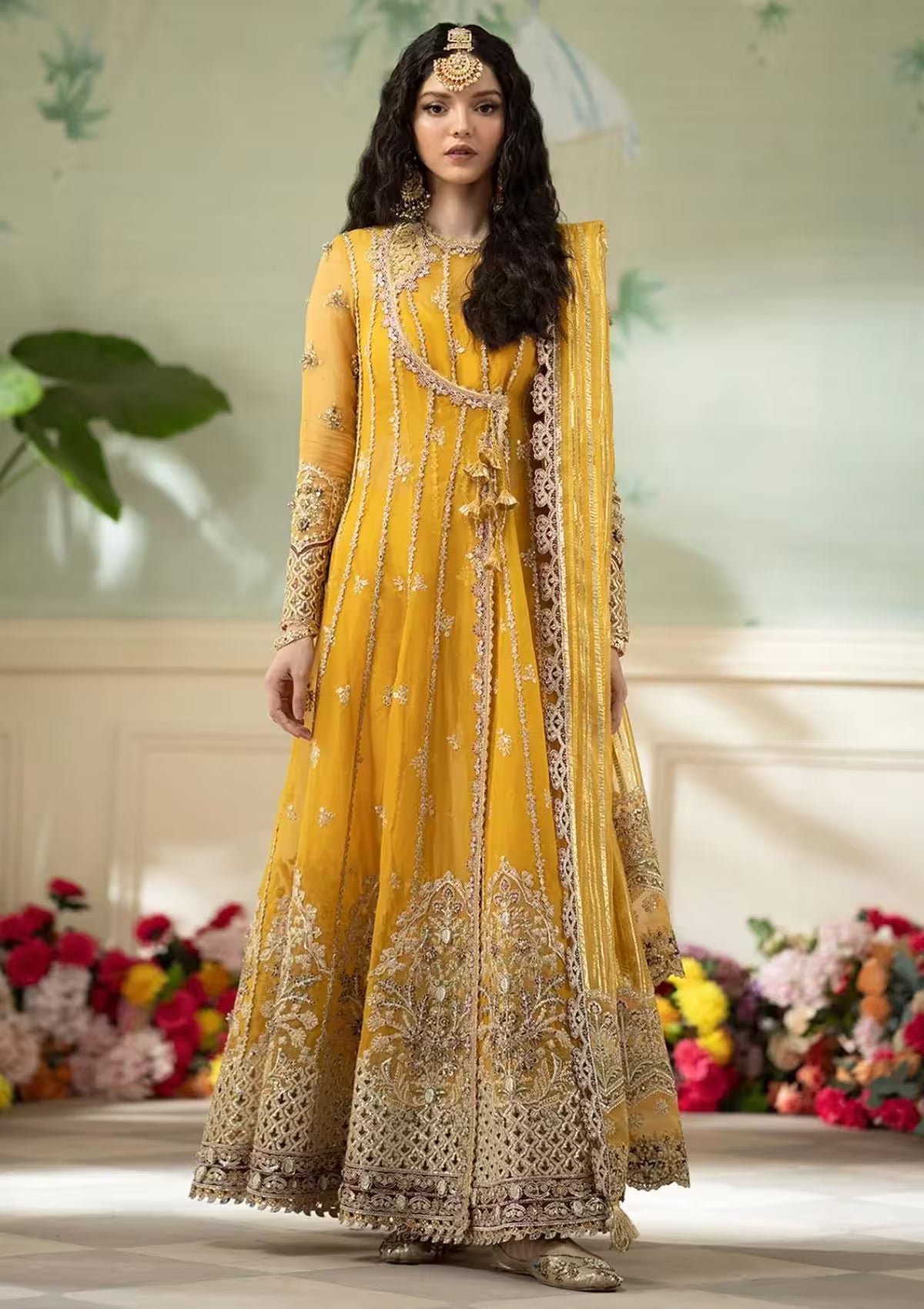 Woman in a yellow embroidered traditional outfit standing in front of floral decorations.