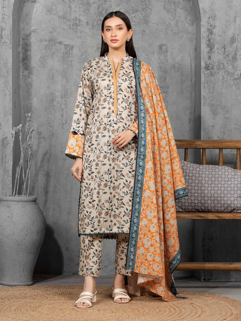 Woman wearing a patterned outfit with an orange dupatta against a gray wall.