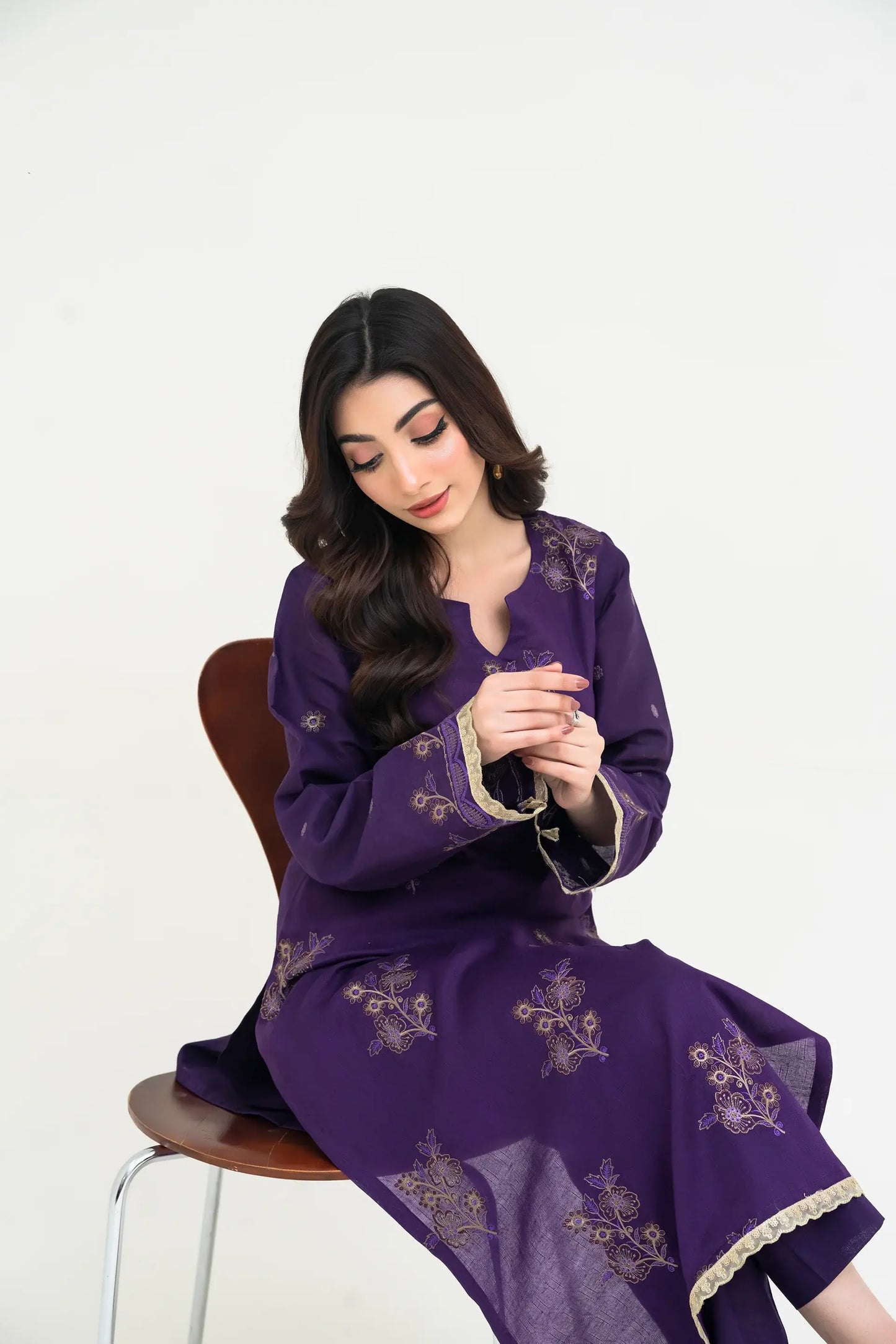 Woman wearing a purple embroidered outfit sitting on a chair against a white background