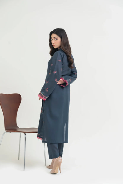 Woman wearing a blue traditional outfit with red patterns, standing next to a chair on a white background.