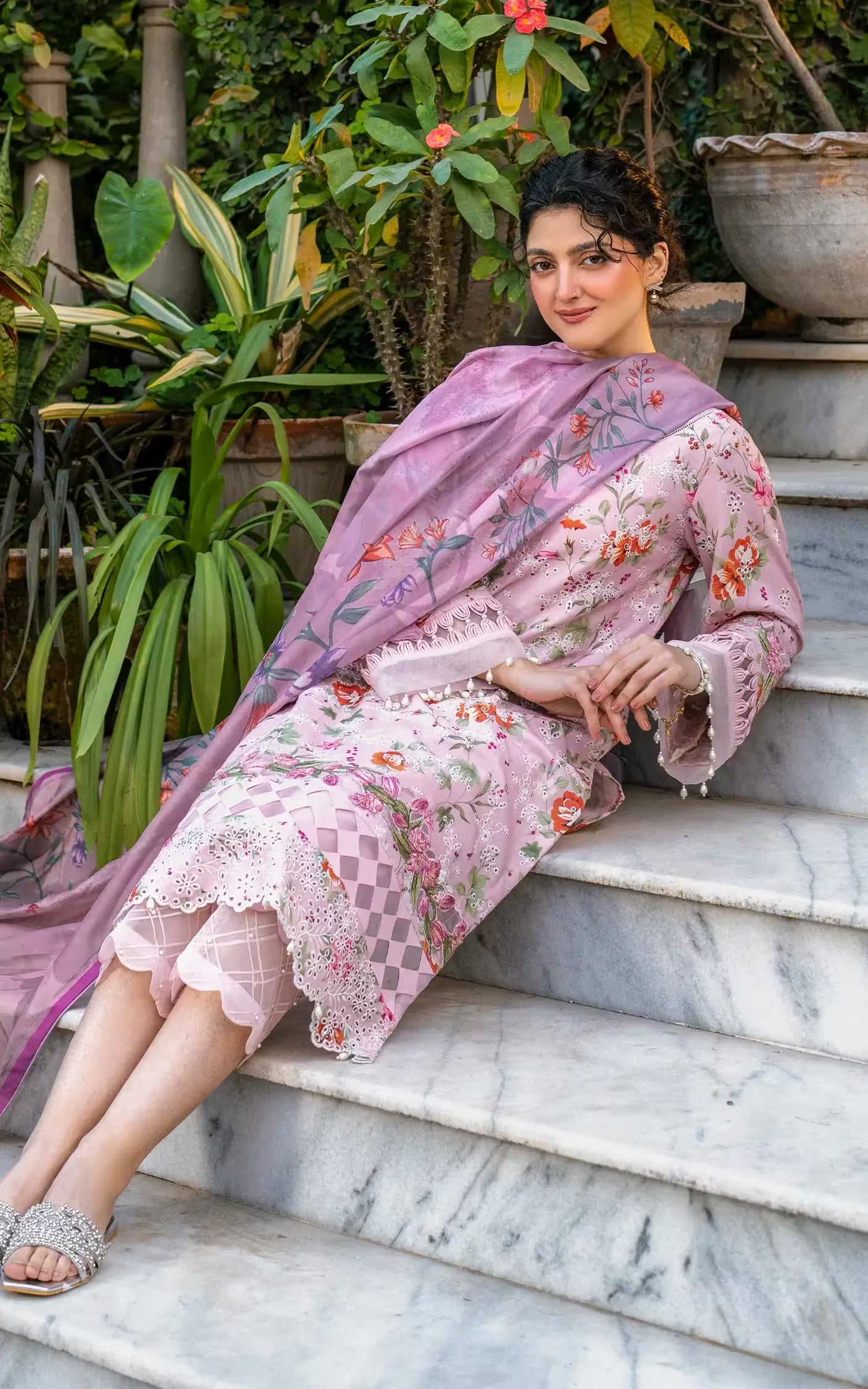 Woman in a floral dress and dupatta sitting on marble steps with plants in the background