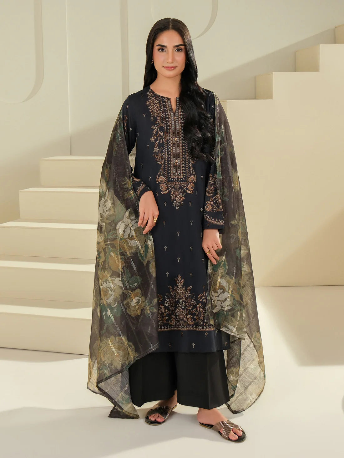 Woman wearing a black embroidered outfit with a sheer green dupatta in a white interior setting.