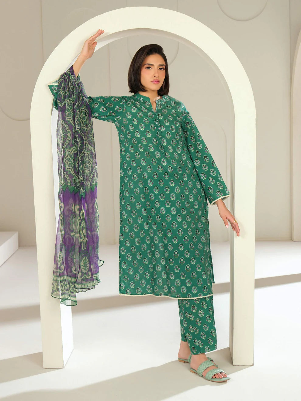 Woman in a green patterned outfit holding a purple scarf, standing in front of a white arch.