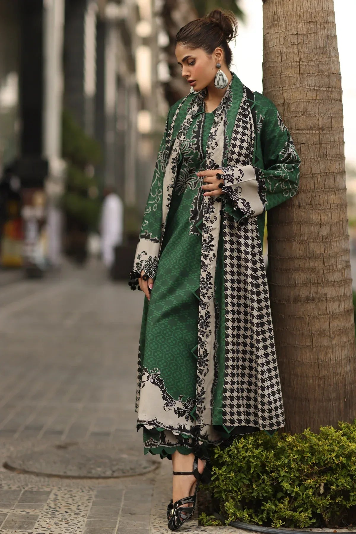 Woman in a green and patterned outfit leaning against a palm tree on a city street.