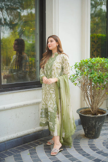 Woman in a green and white traditional outfit standing outside a building with a plant in the foreground.