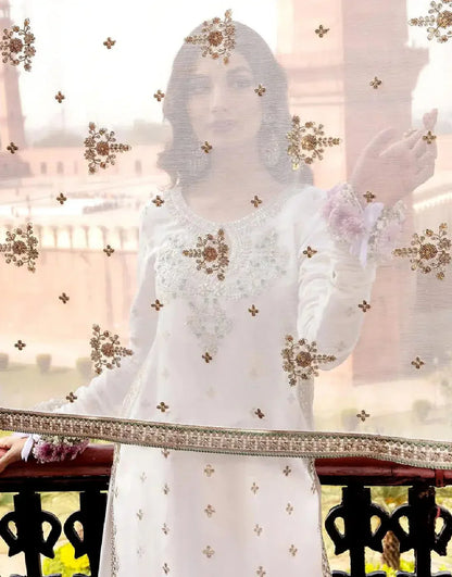 Woman in a white embroidered outfit with gold details, standing in front of a blurred scenic background.