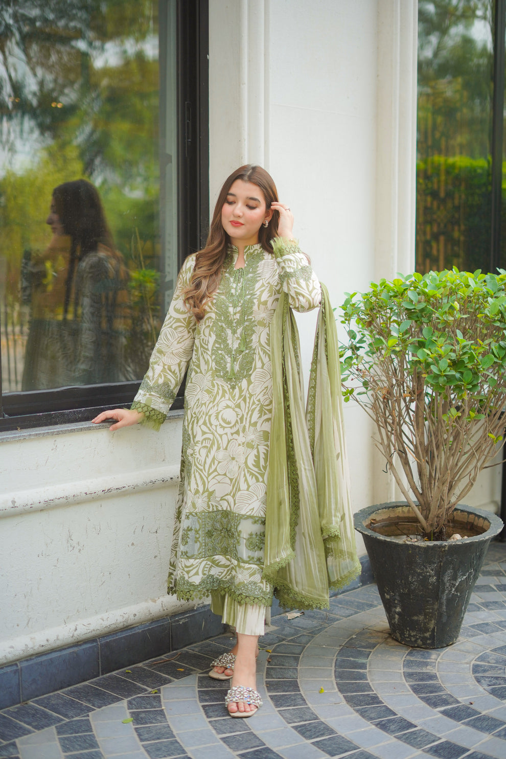 Woman in a green and white traditional outfit standing by a window with a plant in the foreground.