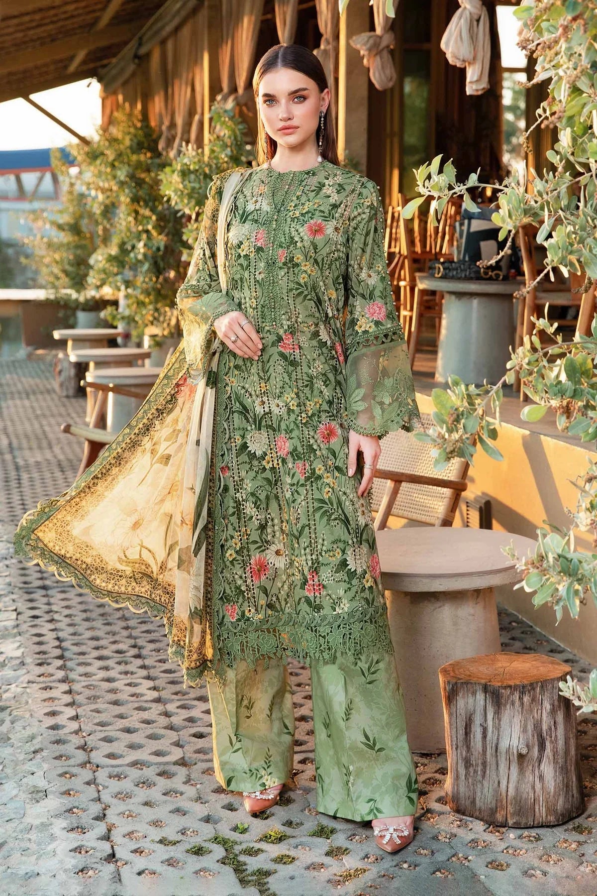 Woman in a green embroidered outfit standing outdoors near wooden furniture and plants.